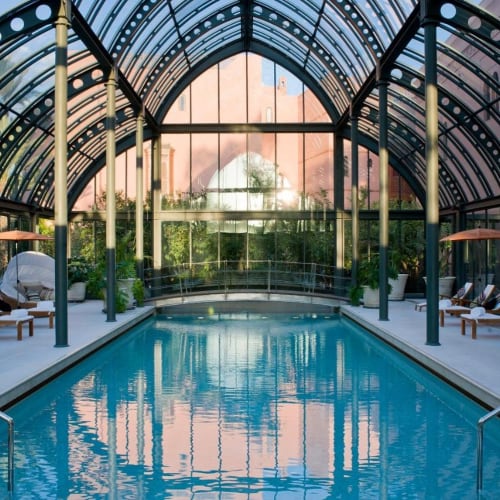 Indoor swimming pool at a luxurious hotel with a glass ceiling and lounge chairs along the sides.