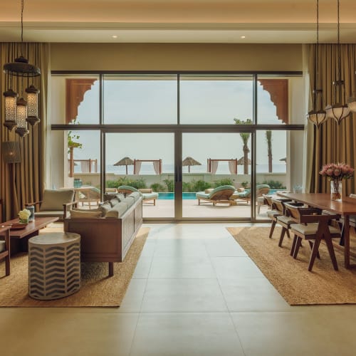 Geräumiges Wohnzimmer einer Strandvilla mit großen Glastüren, die zu einem Poolbereich und Liegestühlen mit Sonnenschirmen im Freien führen, dekoriert mit modernen Holzmöbeln und warmem Licht.