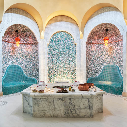 Luxurious Hammam room with ornate turquoise tile benches, intricate tiled walls, arched niches, a central marble platform with spa items, and warm hanging lanterns.
