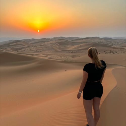 Eine junge Frau in schwarzer Kleidung steht auf Sanddünen bei Sonnenuntergang und blickt auf die weite, rolling Wüstenlandschaft.