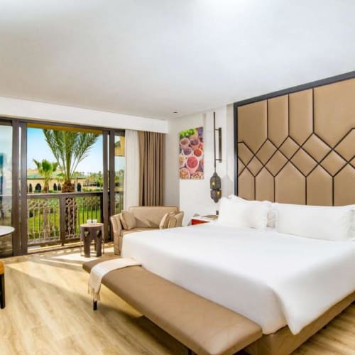 Modern hotel bedroom with a large bed and beige upholstered headboard, two bedside tables with hanging lamps, a small round table with two orange chairs, and balcony doors opening to a resort view with palm trees.