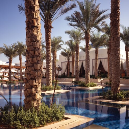 Außenpoolbereich in einem Luxushotel mit hohen Palmen und weißen Gebäuden im Hintergrund bei klarem blauem Himmel.