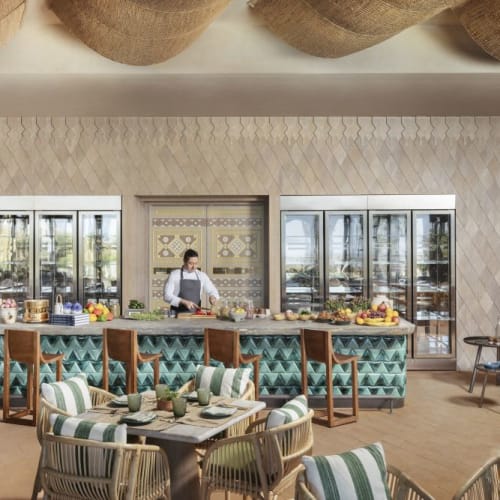 A luxurious restaurant with a relaxed atmosphere, featuring a large turquoise island with a chef preparing meals, surrounded by tables and chairs in a modern setting.