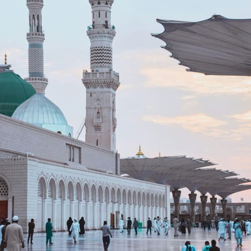Eine weite Ansicht einer Moschee in Medina mit Menschen, großen Sonnenschirmen und Sonnenuntergang im Hintergrund.