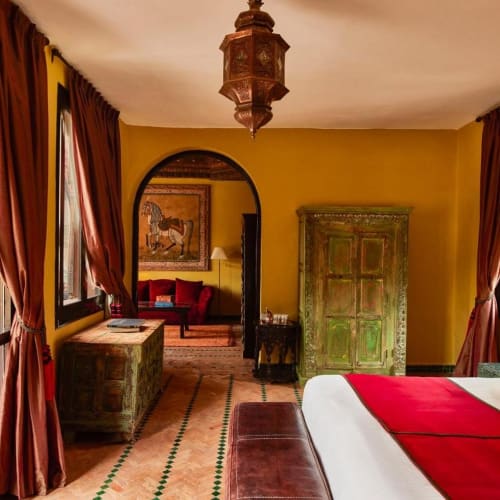 Ein gemütliches Schlafzimmer mit gelben Wänden, großen Fenstern mit braunen Vorhängen und einem Bett mit weißen Bettwäsche und gelben Kissen.