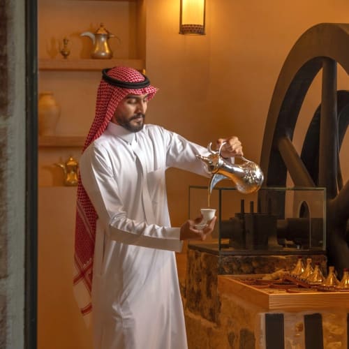 A man in traditional Saudi attire is pouring coffee into a cup inside a cozy, well-lit coffee house with traditional decor.