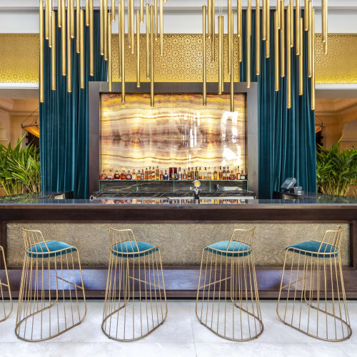 Modern hotel bar with marble counter, six wireframe bar stools with teal cushions, golden hanging pendant lights, and decorative golden and teal curtains in the background.