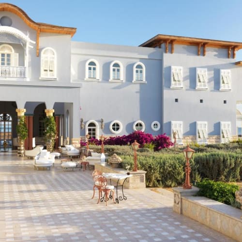 Elegantes mediterranes blaues Hotel mit gefliester Terrasse, Außensitzplätzen, verzierten Säulen und üppiger Begrünung unter klarem blauem Himmel.