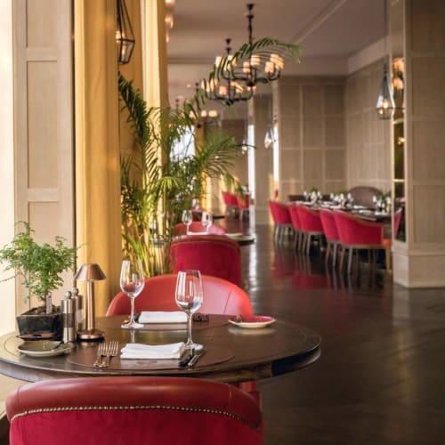 Elegant restaurant dining area with round table set for a meal, featuring red chairs, large plants, and modern pendant lighting.