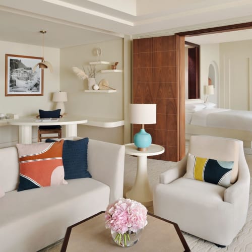 Ein modernes Wohnzimmer mit einem weißen Sofa und Sessel, Dekokissen, einem Couchtisch mit rosa Blumen und einem Blick in ein Schlafzimmer, das durch holzverglaste Schiebetüren getrennt ist.