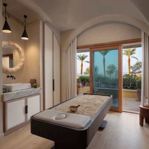 Ein Wellnessraum mit einer Massageliege, einem Waschbecken und einer großen glassglastür, die zu einem Balkon mit Palmen und Meerblick führt.