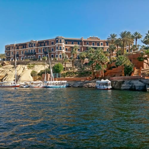 Luxury hotel Sofitel Legend Old Cataract overlooking the Nile River in Aswan, Egypt, with boats docked along the riverbank and palm trees nearby.