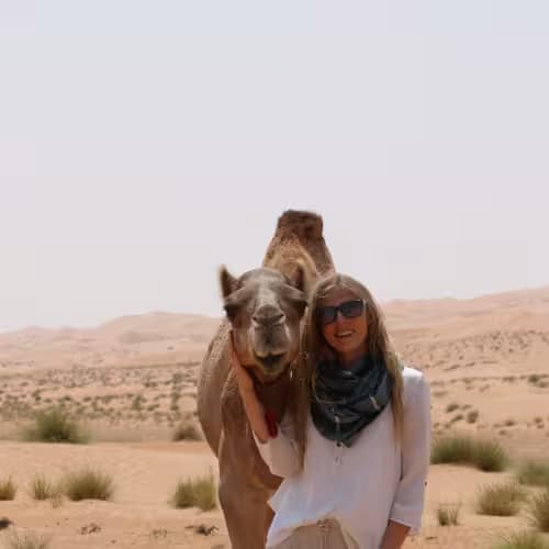 A woman with sunglasses and a scarf standing next to a smiling camel in a desert landscape.