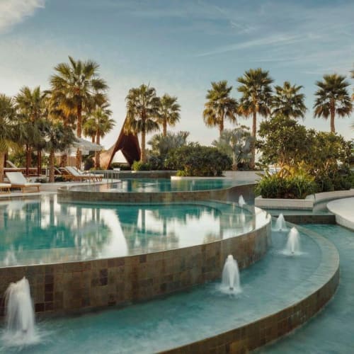 Luxuriöser Resort-Pool umgeben von Palmen und Sonnenliegen unter einem bewölkten Himmel.