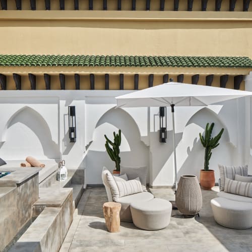 Outdoor terrace at Mazagan Beach Golf Resort featuring plush round chairs, small wooden stools, potted cacti, a white sun umbrella, and a tiled pool with lounge pillows in the background.
