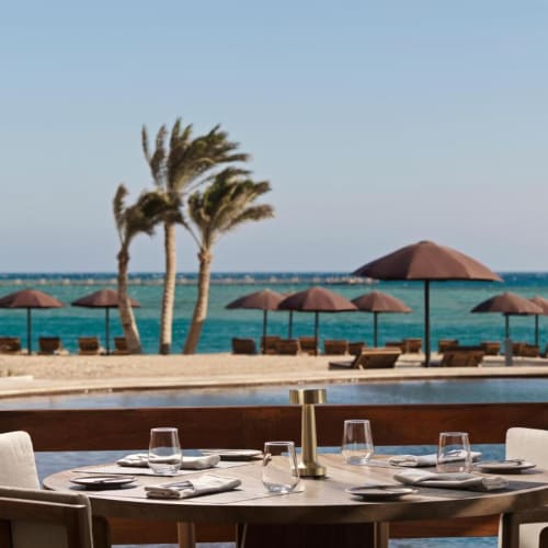 Ein gedeckter Esstisch in einem Restaurant mit Blick auf Palmen, Strandkörbe und das Meer im Hintergrund.