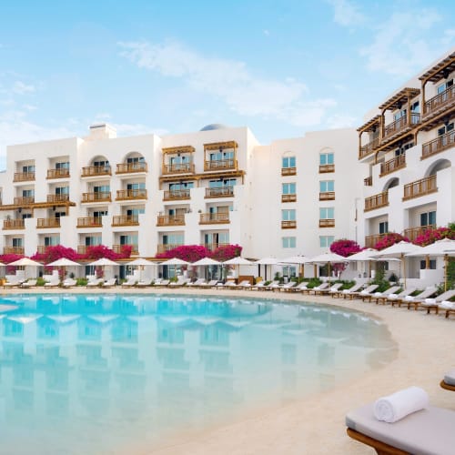 Luxuriöse Hotelfassade mit einem großen, geschwungenen Swimmingpool, umgeben von Liegestühlen, weißen Sonnenschirmen und leuchtend pinken Blumen an einem sonnigen Tag.