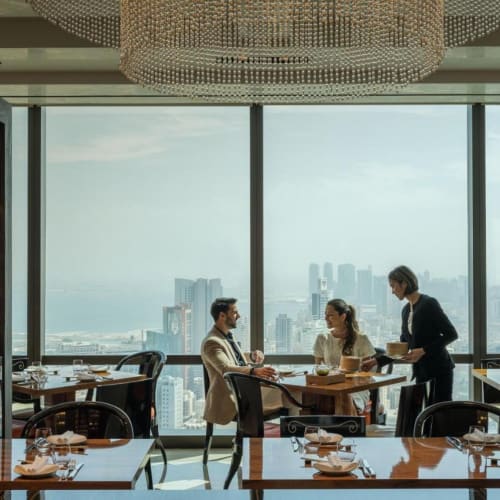 Ein Restaurant mit großen bodentiefen Fenstern, die einen Blick auf die Stadt bieten, mit einem Kellner, der zwei Gäste an einem Tisch bedient.