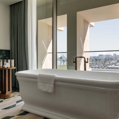 Ein modernes Badezimmer in der The Ned Doha Palace Suite mit einer frei stehenden weißen Badewanne mit einem Handtuch an der Seite, einem hölzernen Beistelltisch mit Toilettenartikeln und großen bodentiefen Fenstern mit Blick auf die Stadt.