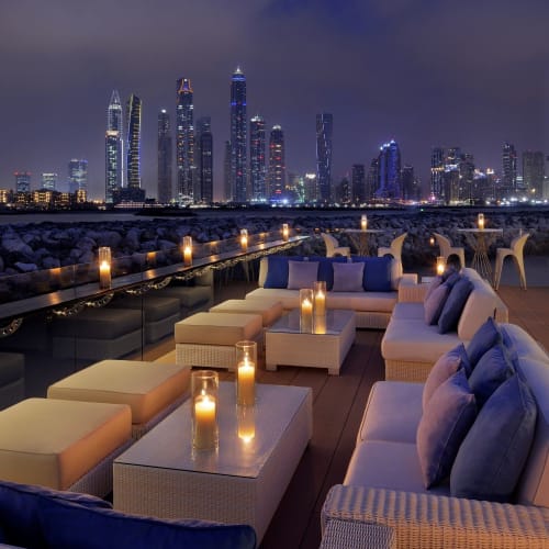 A luxurious rooftop lounge at night with comfortable seating, lit candles, and a view of a modern city skyline with tall skyscrapers.