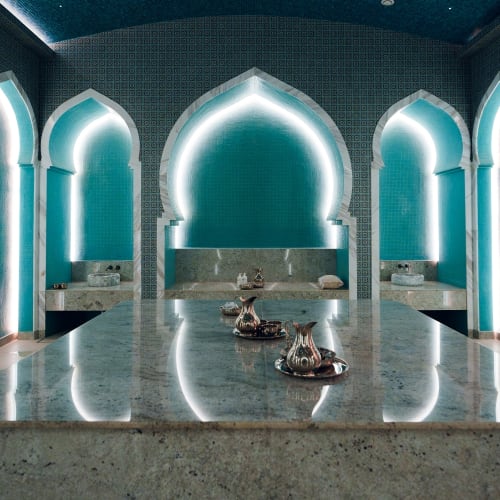 Innenraum eines luxuriösen Hamams mit leuchtend türkisfarbenen, gewölbten Nischen und einem großen zentralen Marmortisch mit silbernen Teekannen.