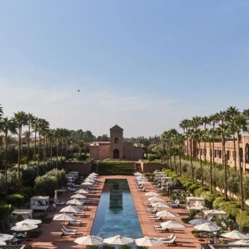 Ein luxuriöser Resort mit einem zentralen Pool, umgeben von Palmen und Sonnenliegen, mit einem Gebäude mit Bögen und einem Turm im Hintergrund.