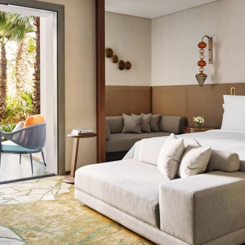 Modernes Deluxe-Hotelzimmer mit King-Size-Bett, beigem Sofa am Fußende und einem Sitzbereich im Freien, der durch eine Glastür mit Palmen im Hintergrund sichtbar ist.