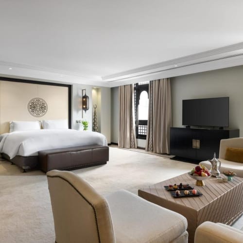 Luxurious hotel suite with a large bed, elegant sitting area with armchairs and a coffee table, and a flat-screen TV.