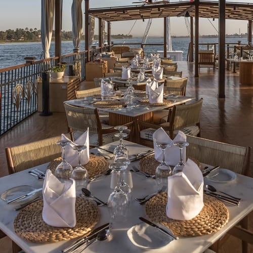 Elegant outdoor dining tables set with folded white napkins, glassware, and cutlery on a sunlit deck overlooking a river with distant greenery.