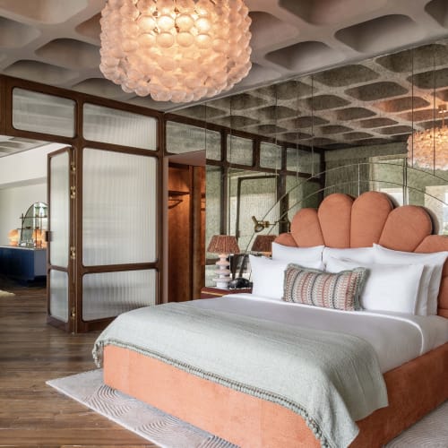 Geräumiges Schlafzimmer in der Park Suite des The Ned Doha mit einem bett mit pfirsichfarbenem muschelförmigem Kopfteil, verspiegelt Wänden, Holzboden und einer einzigartigen Deckenleuchte.