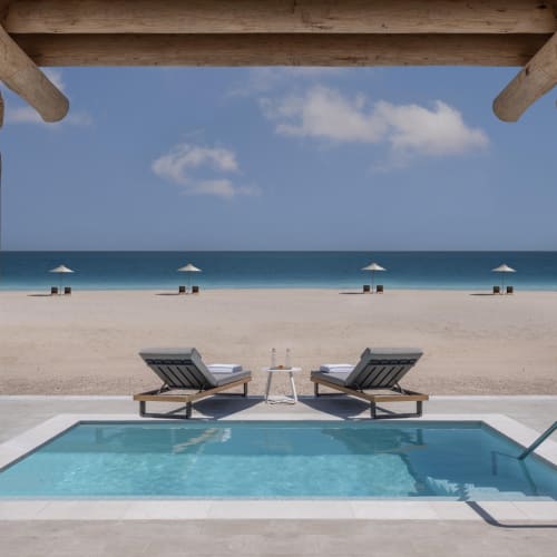 Ein privater Pool mit zwei Liegestühlen und einem kleinen Tisch unter einem Holzpergola, mit Blick auf einen leeren Sandstrand und ruhiges Meer bei klarem blauem Himmel.