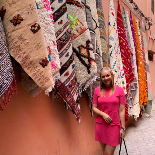 A woman in a pink dress standing in front of colorful embroidered rugs hanging on a pink wall in Morocco.