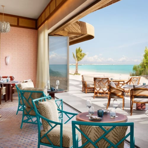 Elegantes Interieur eines Resort-Zimmers am Meer mit großen offenen Schiebetüren, die den Blick auf einen Strand mit Außenbereich und Palmen freigeben.