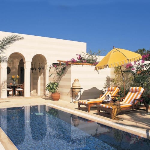 A luxurious poolside area at a beach resort with striped lounge chairs, an umbrella, and lush pink flowering bushes under a clear blue sky.