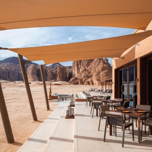 Outdoor dining area at the Banyan Tree AlUla with tables, chairs, and large shade sails in a desert setting with rocky formations in the background.