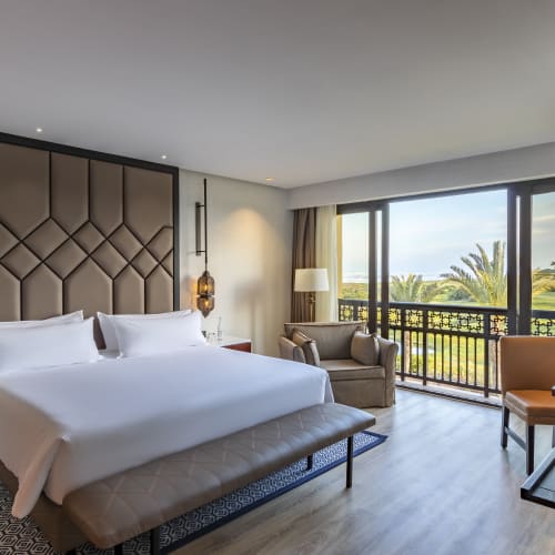 A modern hotel bedroom with a large bed, elegant bedside lamps, seating area, and a sliding glass door opening to a balcony with a view of palm trees and a sunny outdoor landscape.