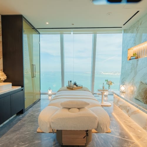 Luxuriöser Spa-Behandlungsraum im Fairmont Doha mit Massageliege und Kissen, Marmorwänden, stimmungsvoller Beleuchtung, Waschbeckenbereich und großen Fenstern mit Meerblick.