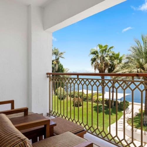 A balcony with wooden chairs overlooking lush gardens, palm trees, and a body of water under a clear blue sky.