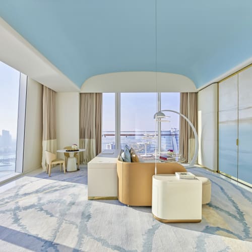 Moderner luxuriöser Wohnraum mit großen bodentiefen Fenstern mit Blick auf eine Stadt und das Wasser, mit blauem Deckenbereich, beigem Sofa, runden Sesseln, kleinem Tisch mit Stühlen und markanten Leuchten.