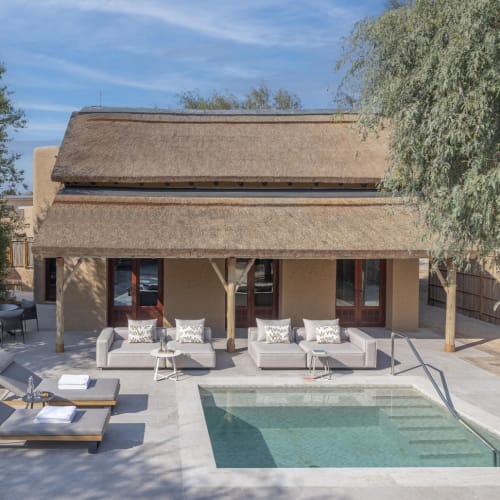 Villa mit Strohdach und Terrasse mit drei modernen Sofas, zwei Liegestühlen mit Handtüchern und einem rechteckigen Pool in einer wüstenähnlichen Außenanlage.