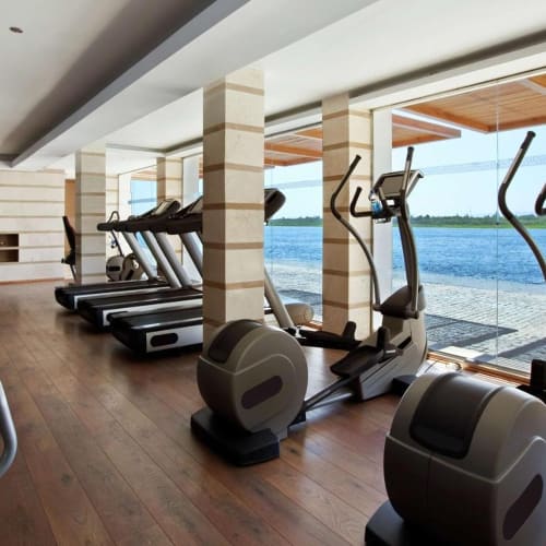 Indoor gym with cardio equipment and large windows overlooking a body of water.