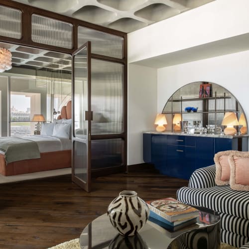 Ein modernes Hotel-Suite-Wohnzimmer mit einem gestreiften Sofa, Glastisch mit Büchern und dekorativer Vase sowie Blick durch geöffnete doppelte Glastüren ins Schlafzimmer.