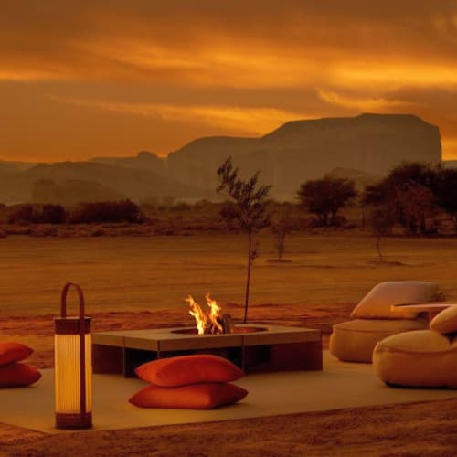 A cozy outdoor seating area with cushions, a fire pit, and a lantern, set against a sunset desert landscape with mountains in the background.