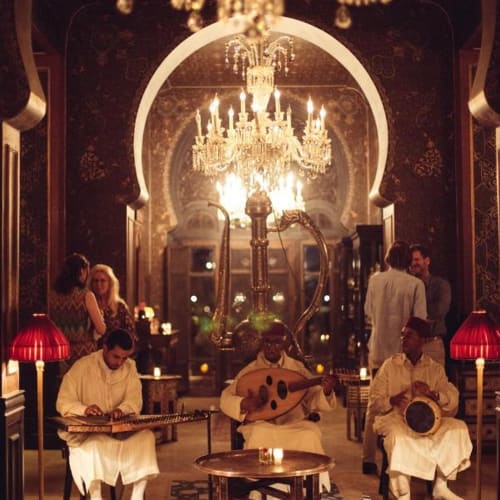 Traditional Moroccan musicians performing in an ornate, warmly lit setting with chandeliers and lush decor.