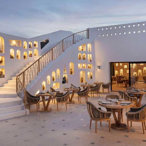Outdoor dining area at a modern hotel at dusk with illuminated wall niches displaying decorative items and wooden tables and chairs arranged on a tiled floor.