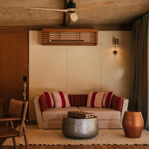 Ein gemütliches Wohnzimmer mit einem weißen Sofa, das mit roten und gemusterten Kissen geschmückt ist, einem runden grauen Hocker, einer geflochtenen Tontopf und einem strukturierten beigen Teppich, mit einer Schiebetür, die den Außenbereich zeigt.