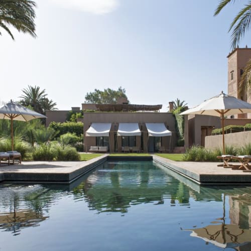 Ein luxuriöser, outdoor Schwimmbecken, umgeben von Liegen, Sonnenschirmen und üppigem Grün, mit einem privaten Haus im Hintergrund.