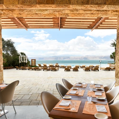Dining area inside a restaurant overlooking a pool and the Dead Sea with lounge chairs in the background.