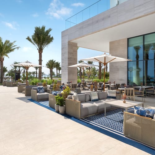 Luxurious outdoor restaurant terrace at a beach resort with palm trees, umbrellas, cushioned seating, and ocean view.