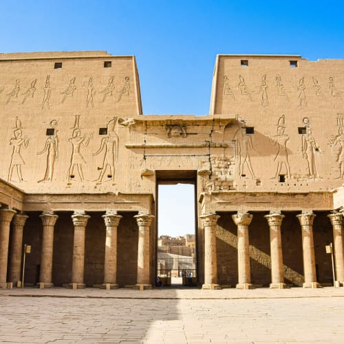Der Tempel von Edfu mit detaillierten Reliefs von altägyptischen Figuren, umgeben von kolonnenartigen Strukturen unter einem klaren blauen Himmel.